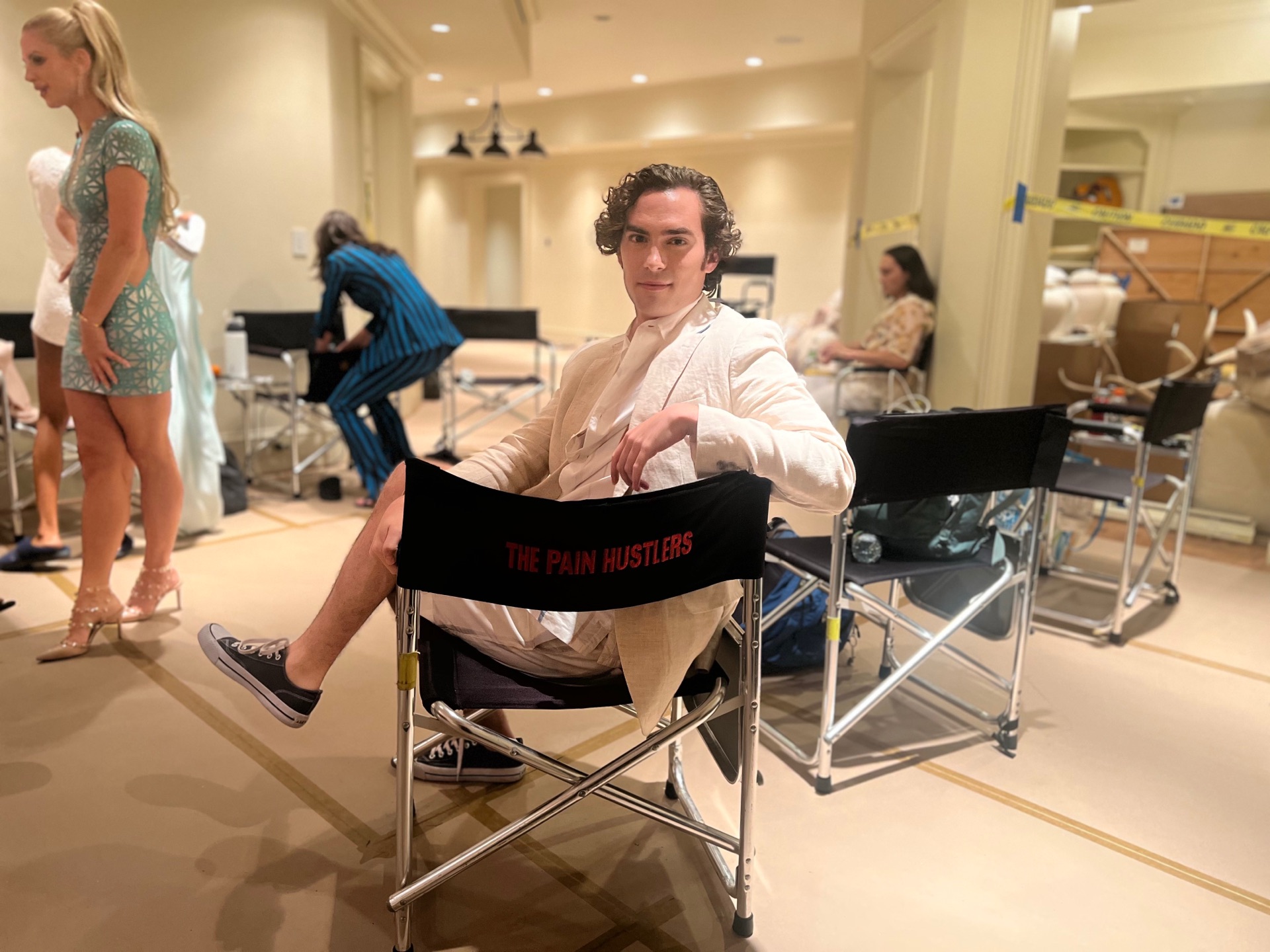 Neil in cream suit sitting coolly in a director's chair stamped "THE PAIN HUSTLERS," background actors milling behind him — actors' holding area, landscape, face visible